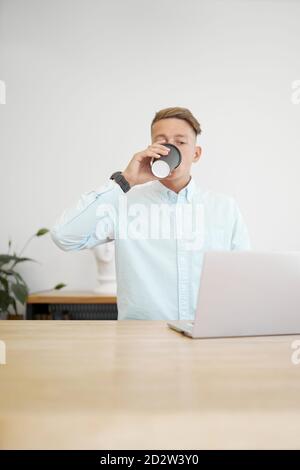 Busy male freelancer sitting at wooden table drinking from a takeaway coffee and working on remote project while browsing netbook Stock Photo