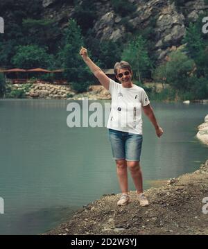 Happy elderly woman by the lake with hand in the air. Senior woman with expression of achievement. Stock Photo