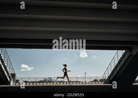 Side view of determined female athlete running fast during outdoor workout near stairways of stadium Stock Photo
