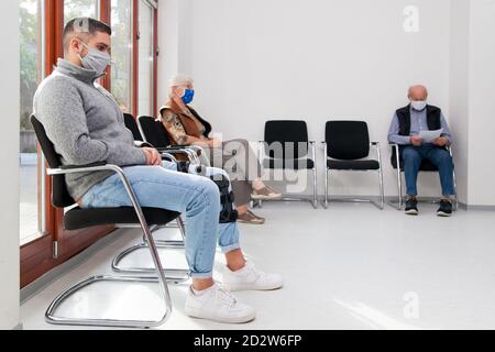 Young and old people with face masks keeping social distance in a waiting room of a hospital or office -  focus on the young man in the foreground Stock Photo