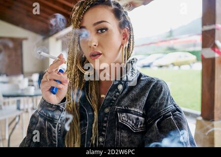 Confident female with extraordinary appearance sitting in outdoors cafe and smoking cigarette while looking at camera Stock Photo