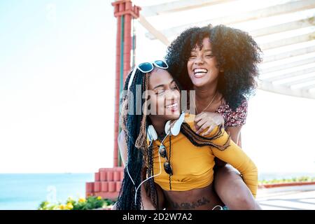 Cheerful ethnic Woman with curly hair piggybacking delighted female friend with braids while having fun on promenade at weekend in summer