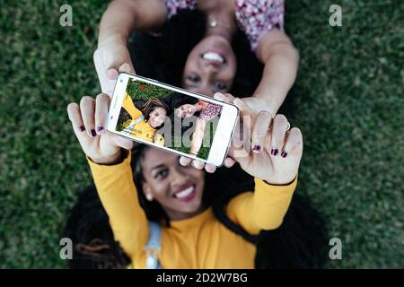 Top view of cheerful female best friends lying on green grass together and taking photo on smartphone camera while enjoying weekend Stock Photo