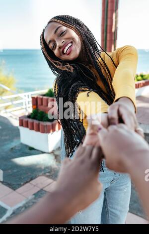 Delighted African American female with braids holding hands of crop friend while standing on embankment in summer and looking at camera