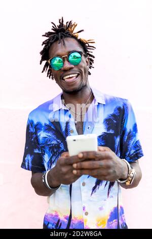 Cheerful trendy African American male in shirt with tropical print and with dreadlocks standing near pink building and chatting on social media via mobile phone Stock Photo