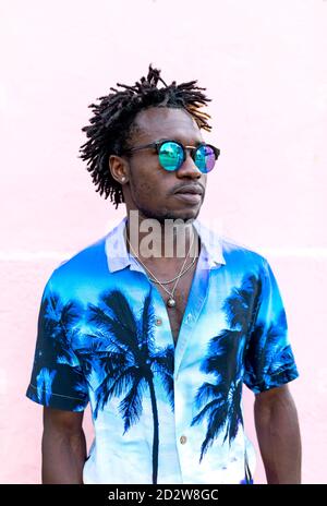Confident African American male wearing trendy shirt with palm trees and stylish sunglasses standing on street near building with pink wall Stock Photo