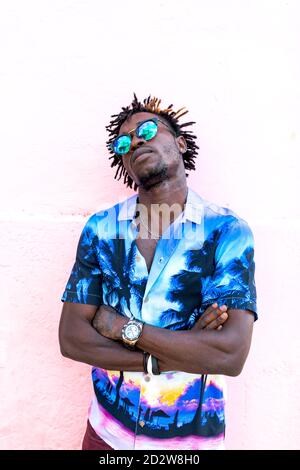Confident African American male wearing trendy shirt with palm trees and stylish sunglasses standing with arms crossed on street near building with pink wall Stock Photo