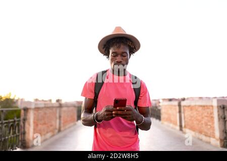 Handsome African American male traveler with backpack standing on bridge and using smartphone Stock Photo
