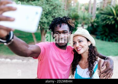 Cheerful multiracial traveling couple hugging and taking selfie on smartphone while standing in green tropical park during summer vacation Stock Photo