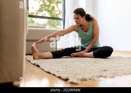 Peaceful female in sportswear practicing yoga in Seated Forward Bend pose and stretching legs while sitting on carpet in living room Stock Photo