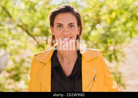 Charming female in leather jacket standing in park in fall and looking at camera Stock Photo