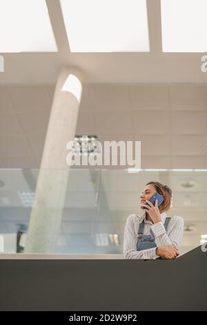 From below of busy female employee standing in contemporary office lounge and speaking about project while working and looking away Stock Photo