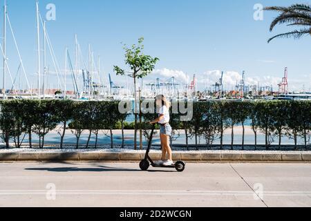 Full body side view of young female in casual outfit riding electric scooter on paved road near urban embankment in sunny summer day Stock Photo