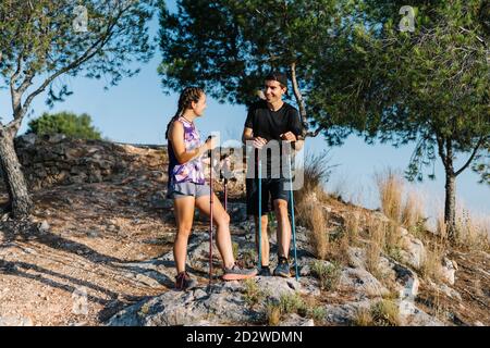 Low angle of happy young man and Woman in activewear with trekking poles having break while standing on mountain slope during hiking and trail running together in summer day in nature Stock Photo