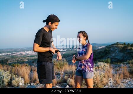 Side view of happy young man and Woman in activewear checking their activity wristband after trail running together in mountainous terrain Stock Photo