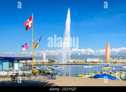 The Swiss flag is flying on the shore of the Lake Geneva in Geneva, Switzerland, on a sunny morning with the water jet fountain in the distance. Stock Photo