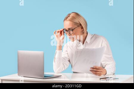 Female office employee looking through documents, making report, doing paperwork near laptop at desk on blue background Stock Photo