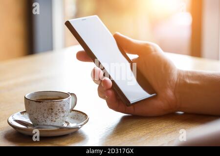 Close up of man's hands holding mobile smart phone with blank copy space screen. Image of man sitting at a table with a coffee in cafe and using mobil Stock Photo
