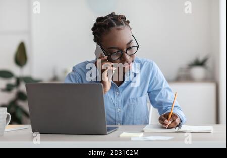 Black Female Office Worker Talking On Cellphone With Client And Taking Notes Stock Photo