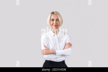 Positive blonde business lady in office wear crossing her arms and looking at camera over light grey studio background Stock Photo