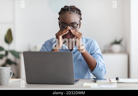 Eye Strain. Tired African Female Office Worker Sitting At Workplace Massaging Nosebridge Stock Photo