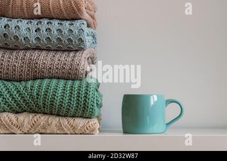 Stack of warm knitted women's sweaters and cup of hot drink on white background. Autumn or winter concept Stock Photo