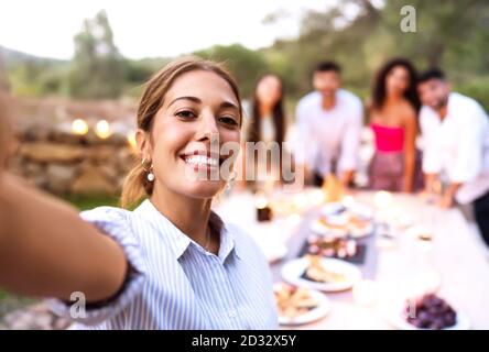 Cute smiling young woman make a self-portrait of herself and friends in the background celebrating in the garden at sunset - Happy friends moment of a Stock Photo