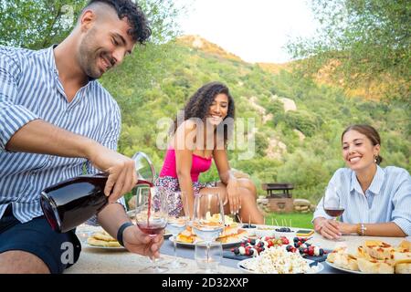 Group of three multiethnic friends celebrating outdoor - Selective focus on the young man with a striped shirt pouring red wine into the glass to his Stock Photo