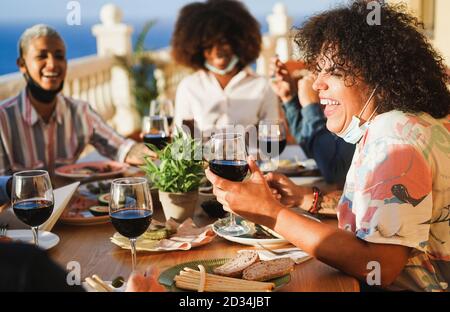 Young multiracial people eating and drinking red wine while wearing protective masks - Happy friends having fun doing dinner at restaurant patio - Soc Stock Photo