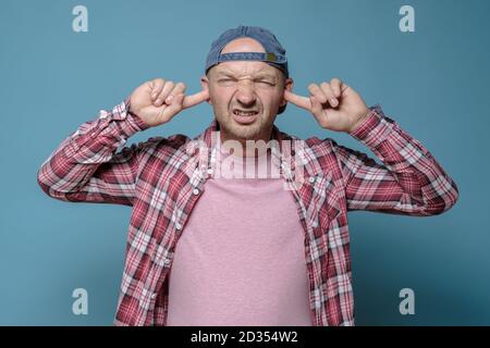 Disgruntled man covered ears with fingers, he was tired of the noise and unpleasant sound, bares teeth and squints eyes.  Stock Photo