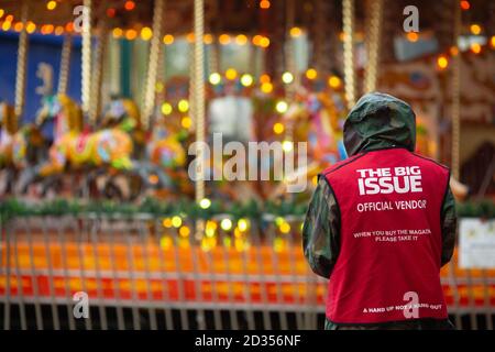 A Big Issue seller stands near the carousel ride near Bournemouth Pier the November wind and rain, Dorset. 13 November 2014. Photo: Neil Turner Stock Photo