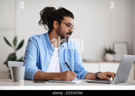 Indian male entrepreneur using laptop and taking notes while working in office Stock Photo