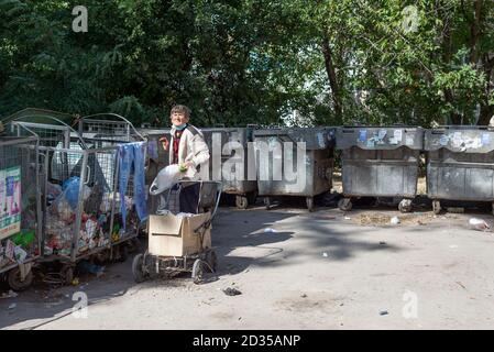 old homeless woman begging through the trash heaps, standing at the dumpster with waste in the landfill Feeling cold. Portrait of a cheerless aged wom Stock Photo