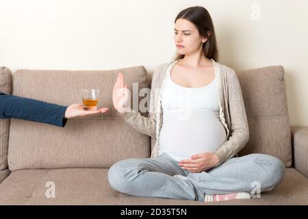 Pregnant woman sitting on the sofa refuses to drink whisky and makes stop gesture to the glass. No alcohol during pregnancy concept. Stock Photo