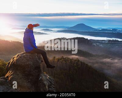 Hiker in blue jacket sit alone on the rock summit. Wonderful daybreak view to heavy mist in deep valley. Man shadowing eyes