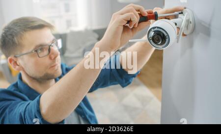 Young Man in Glasses Wearing a Blue Shirt is Screwing a Modern Wi-Fi Surveillance Camera with Two Antennas on a White Wall at Home. Stock Photo