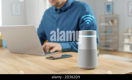 Close Up Shot of a Modern Silver Bluetooth Speaker With Digital Wi-Fi Icon Over It. Man in the Background Sits at the Table and Works on His Laptop Stock Photo