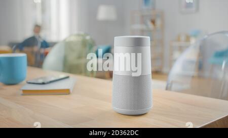 Close Up Shot of a Modern Silver Wireless Speaker Standing on a Table at Home. Man in the Background Sitting on the Sofa. Stock Photo