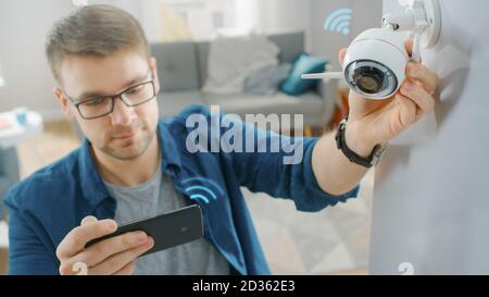 Young Man in Glasses Wearing a Blue Shirt is Adjusting a Modern Wi-Fi Surveillance Camera with Two Antennas on a White Wall at Home. He's Checking the Stock Photo