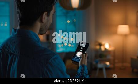 Young Handsome Man is Holding Smartphone with Active Smart Home Application. Person is Tapping the Screen to Turn On Off the Lights in the Room. It's Stock Photo