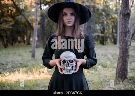 Young beautiful cute woman in dark dress and witch's hat holds a skull in her hands. Halloween party costume. Forest, park with autumn trees Stock Photo