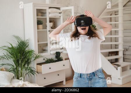 Young woman in jeans and white T-shirt wearing virtual reality helmet plays game, works, watches video in room next to bed. Concept of modern technolo Stock Photo