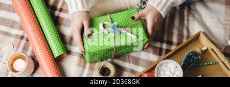 Young woman sits on plaid in cozy knitted woolen white sweater, socks and wraps Christmas gift in polka dot wrapping paper. Wooden tray with mug of co Stock Photo