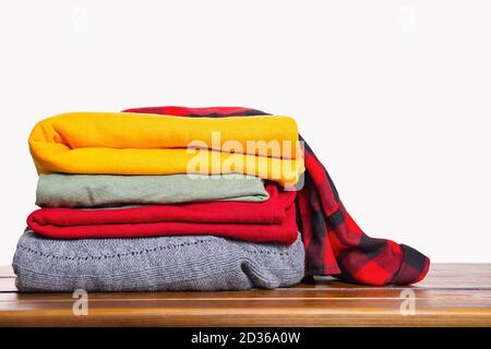 A stack of folded autumn winter clothes on a wooden table on a white background. Clothes in bright trendy colors. Cozy warm knitted sweaters and a che Stock Photo