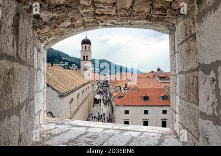 View from stone window of Dubrovnik city center, old tower and crowd. Crowded touristic narrow street in old town, View of red rooftops and tower. Stock Photo