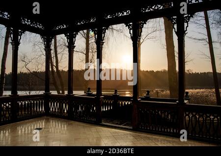 View of sunrise on the lake in spring from wooden openwork gallery or gazebo. Forest silhouettes and the rays of the rising sun Stock Photo