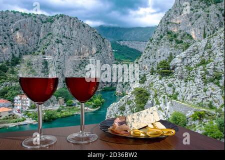 Two glasses of wine with cheese and meat snacks with view of Cetina River, mountains and old town Omis, Croatia. Cetina river canyon in Dalmatia regio Stock Photo