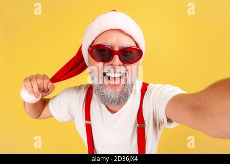 Closeup positive smiling gray bearded man in sunglasses and santa claus hat looking at camera with toothy smile, posing making selfie. Indoor studio s Stock Photo