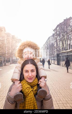 Happy mother carrying daughter on her shoulders in outdoor city street scene in winter, Vitosha Boulevard, Sofia, Bulgaria Stock Photo