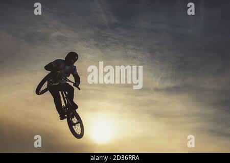 Urban biker performing acrobatic jump at sunny sky - Guy riding bmx bicycle at extreme sport competition on sunny afternoon - Alternative lifestyle co Stock Photo